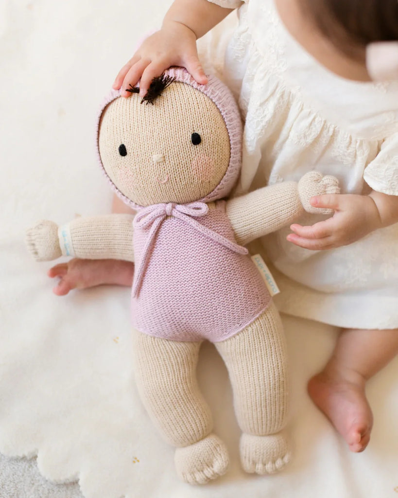 A child in a white dress sits on a soft surface, gently holding the Cuddle + Kind My First Baby (Lavender), a fair trade hand-knit doll dressed in a pink hooded outfit—ships in one week.