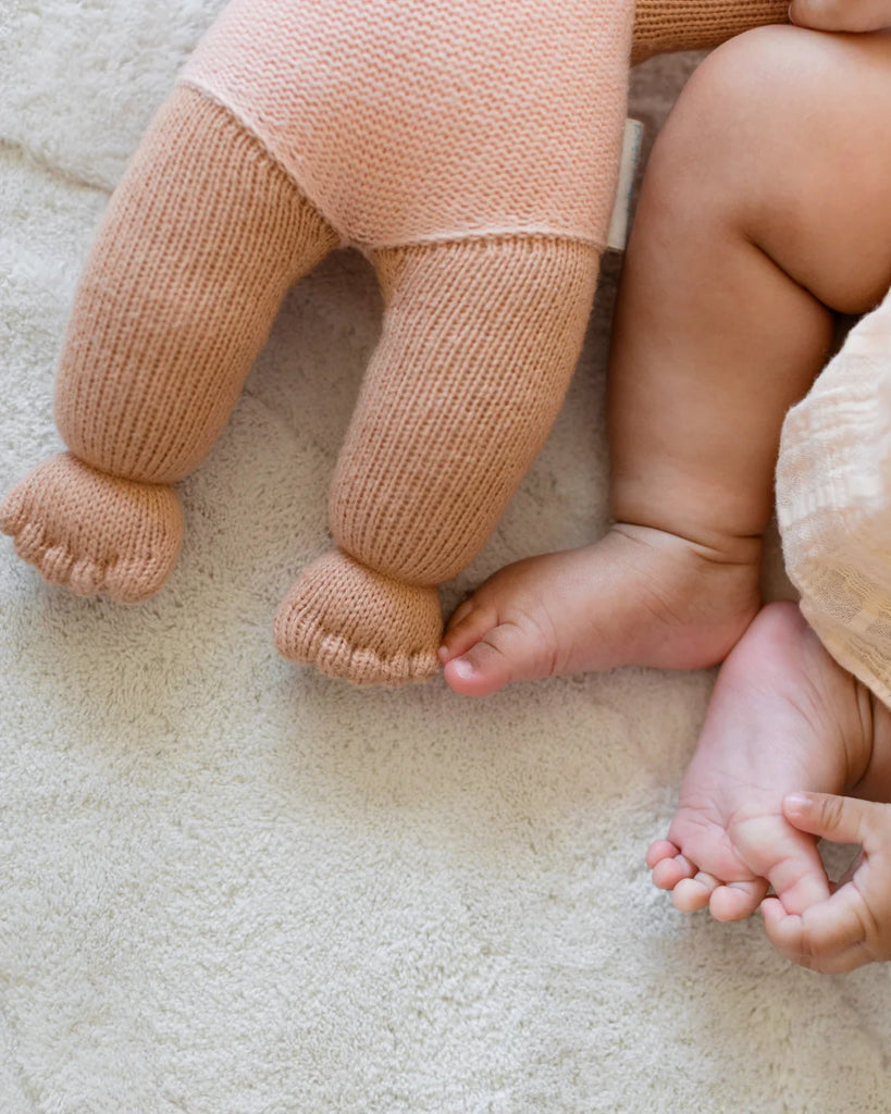 A close-up of a baby’s bare feet beside the Cuddle + Kind My First Baby (Peach), a soft, hand-knit doll made from natural cotton yarn, both resting on a cream-colored blanket. Ships in one week.