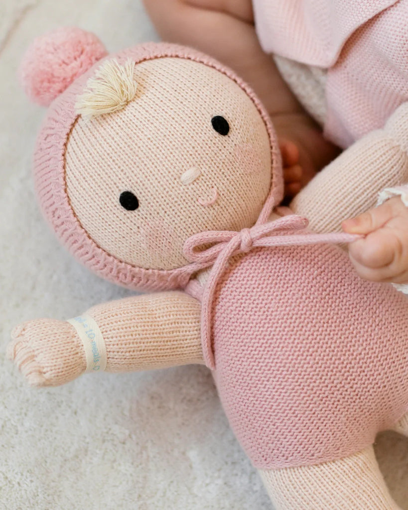 The Cuddle + Kind My First Baby (Rose) is a soft, hand-knit baby doll in a pink hooded outfit with a pom-pom, made from natural cotton yarn. It features a gentle face and wristband, and ships in one week.