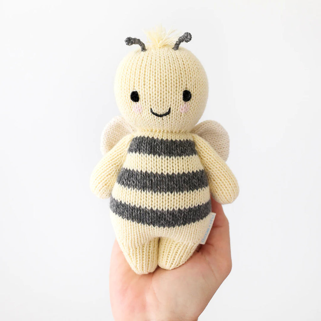 A hand holding a Cuddle + Kind Baby Bee plush toy with black and grey stripes and smiling face against a white background, crafted from Peruvian cotton yarn.