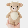 A hand holds a Cuddle + Kind Baby Cow plush toy, shaped like a light brown cow with soft Peruvian cotton yarn. It features small beige horns, a white snout and belly, black eyes, and a gentle expression on a plain white background.