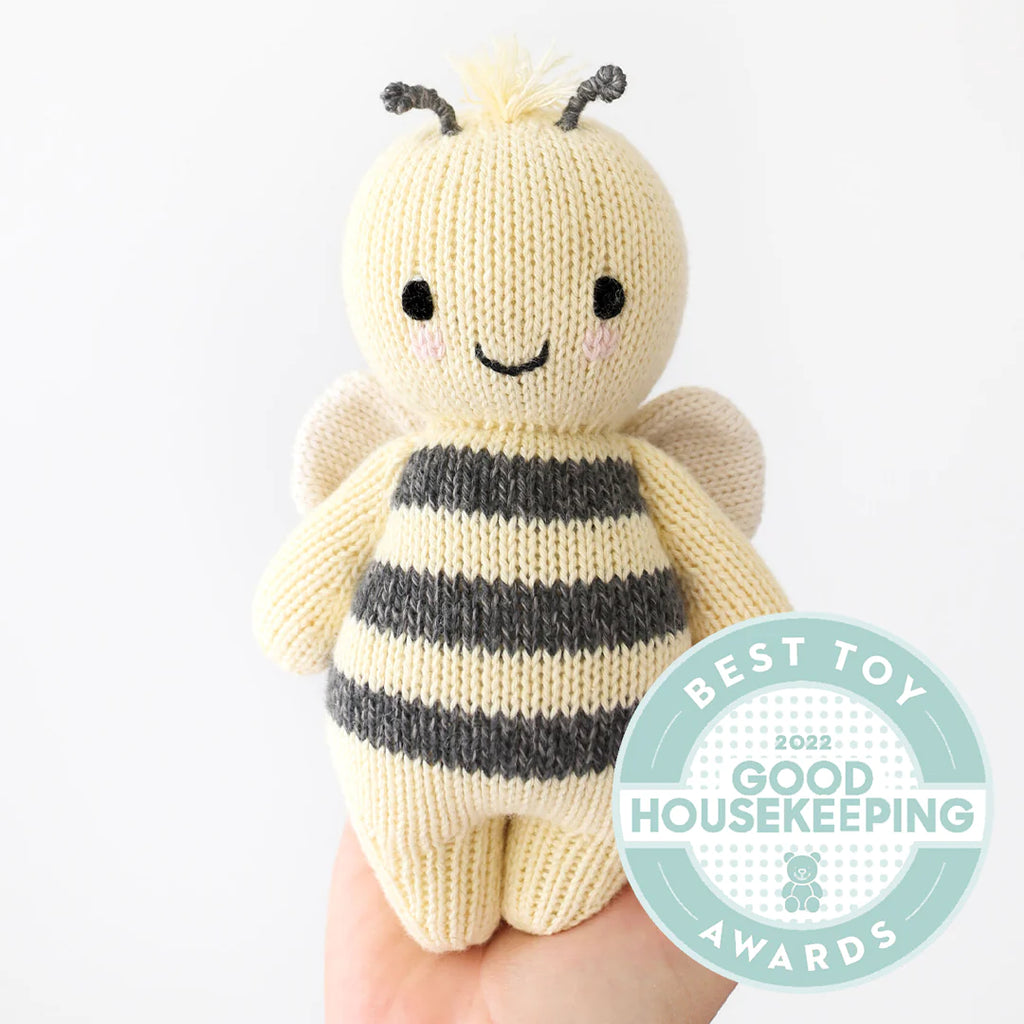 Hand holding a Cuddle + Kind Baby Bee with a smiling face, featuring yellow and black stripes and antennae, against a white background. A badge on the image states "Best Toy Good".