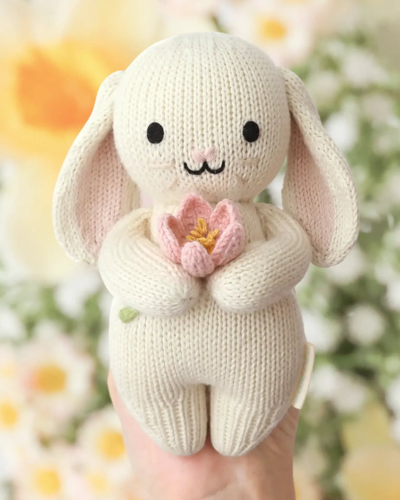 A hand holds the Cuddle + Kind Baby Bunny (Blooming Tulip), an heirloom-quality, hand-knit bunny with long ears, black eyes, a stitched smile, and a pink flower, crafted from soft Peruvian cotton yarn. Blurred floral background.