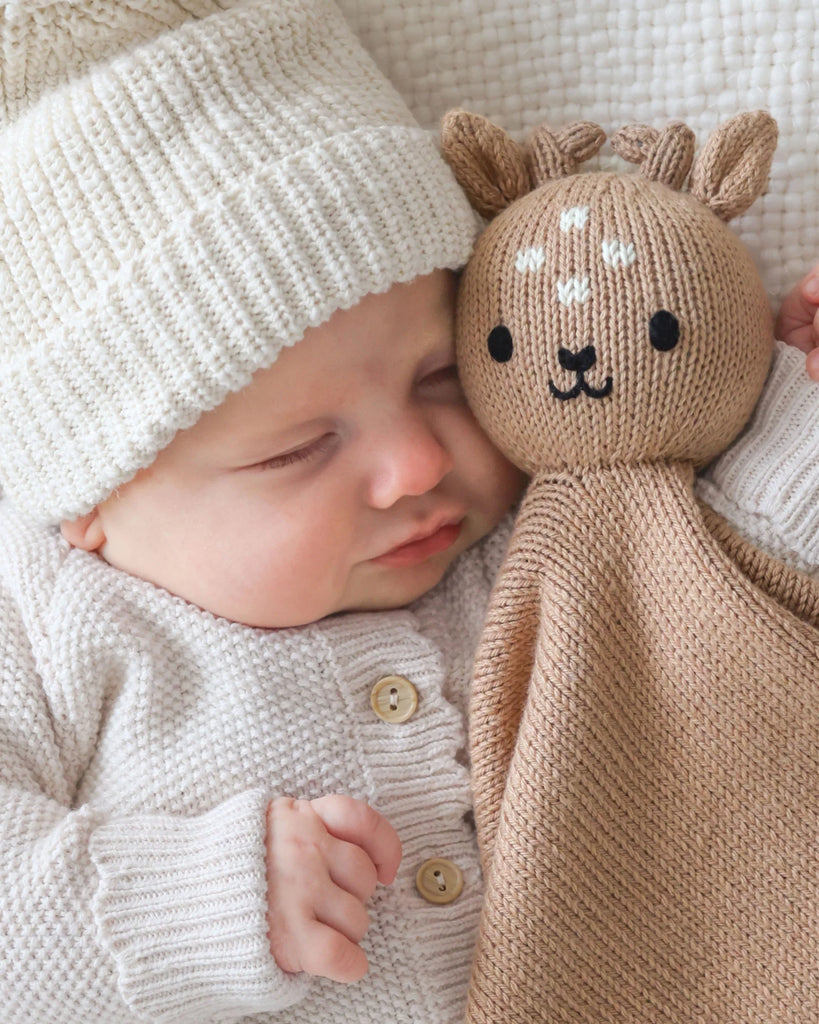 A baby in a cream knit sweater and hat sleeps while cuddling the Cuddle + Kind Organic Cotton Baby Fawn Lovey, an heirloom-quality plush deer toy with embroidered details.