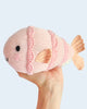 A hand holds a pink Cuddle + Kind Baby Fish plush with embroidered eyes, a smiling mouth, and detailed fins and scales against a light blue background.