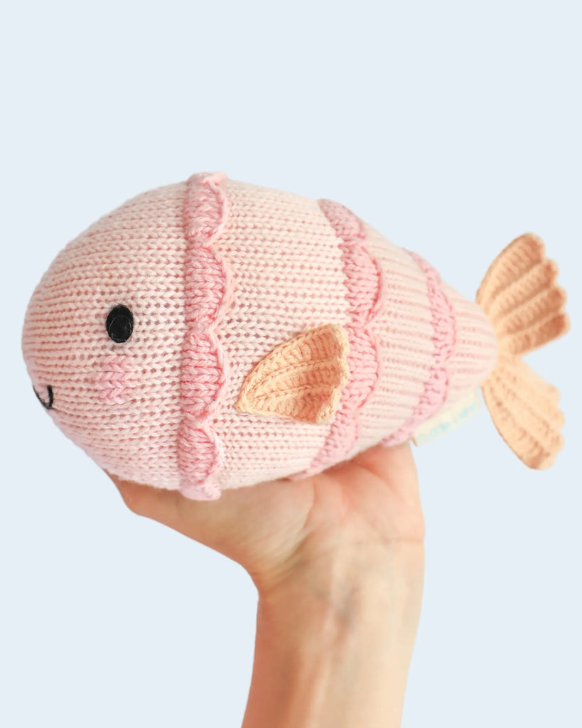 A hand holds a pink Cuddle + Kind Baby Fish plush with embroidered eyes, a smiling mouth, and detailed fins and scales against a light blue background.