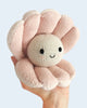 A hand holds a Cuddle + Kind Baby Pearl, a hand-knit plush clam with a round cream face, soft pink shell, black eyes, and a small stitched smile, made from natural cotton yarn.