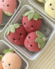 Three Cuddle + Kind Baby Strawberry plush toys, hand-knit from soft Peruvian cotton yarn with smiling faces and green leafy tops, sit in a white container on a yellow striped surface. Heirloom-quality plush fruit toys are partially visible nearby.