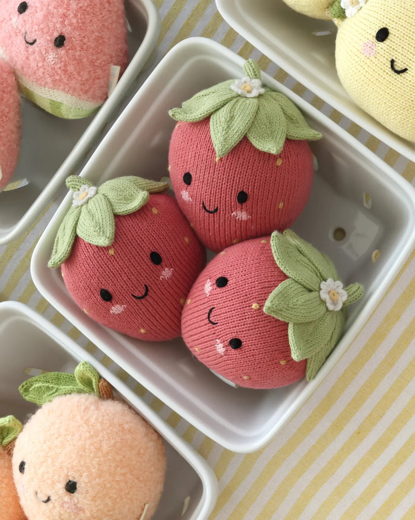 Three Cuddle + Kind Baby Strawberry plush toys, hand-knit from soft Peruvian cotton yarn with smiling faces and green leafy tops, sit in a white container on a yellow striped surface. Heirloom-quality plush fruit toys are partially visible nearby.