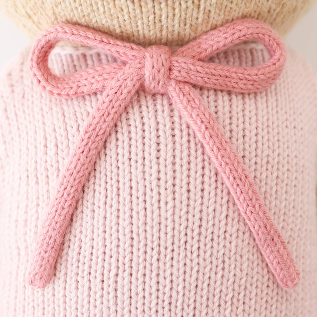 Close-up of a pink knitted bow on Cuddle + Kind Bella the Honey Bear, a pale pink hand-knit doll. The detailed stitches highlight the sustainable, fair trade craftsmanship.