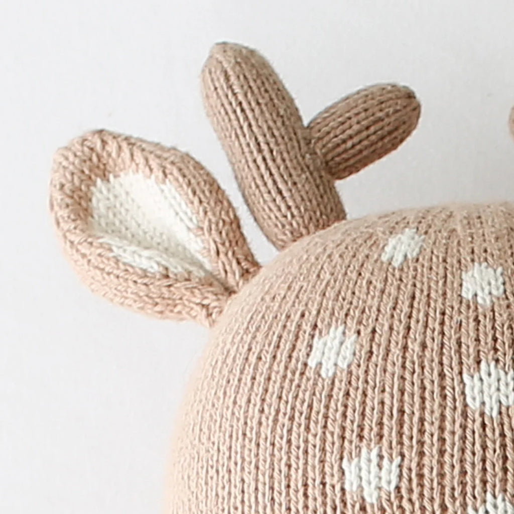 Close-up of a beige hand-knit Cuddle + Kind Elliott the Fawn doll hat with white spots, a soft deer ear, and two antlers on top—crafted from sustainable fair trade materials.