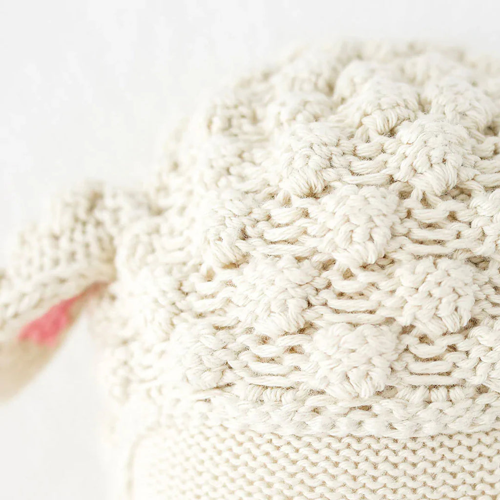 Close-up of Cuddle + Kind Avery the Lamb’s cream-colored, 100% cotton yarn fabric, featuring a soft, bumpy knit texture and a cozy look. Part of the hand-knit doll's fabric is tied in a knot to the left.