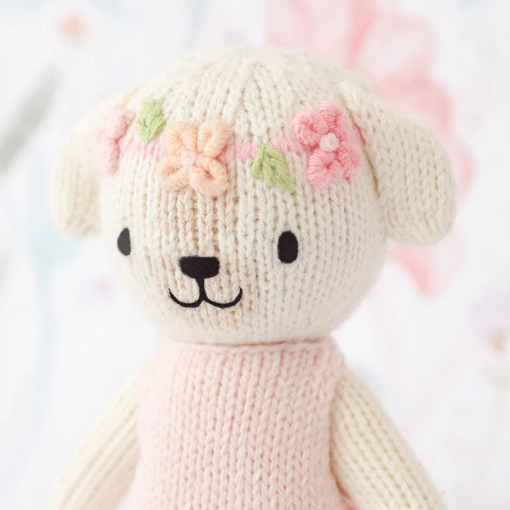 A Cuddle + Kind Tiny Charlotte The Dog with a light pink sweater. The sustainable toy has a white face, a subtle smile, black nose and eyes, and a crown of delicately stitched pink and orange flowers.