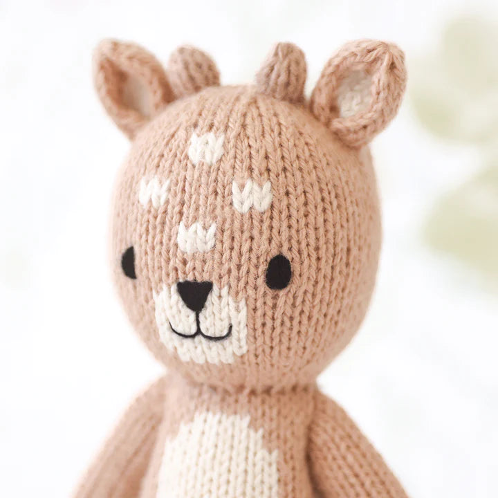A close-up of a Cuddle + Kind Tiny Elliott The Fawn toy depicting a cute deer with a gentle expression, featuring white spots on its forehead and small, pointed ears. The toy is made of soft brown and white yarn.
