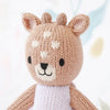 A cute, hand-knit stuffed animal resembling Tiny Violet The Fawn, with a smiling face, prominent ears, and heart-shaped spots on its forehead. It has a soft pink and brown color scheme.