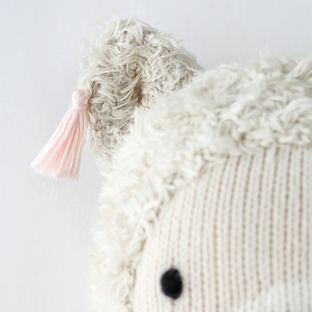 Close-up of Cuddle + Kind Lola the Llama, a hypoallergenic stuffed animal with cream textured fur, black stitched eye, and a fluffy ear accented by a light pink tassel, set against a plain white background.