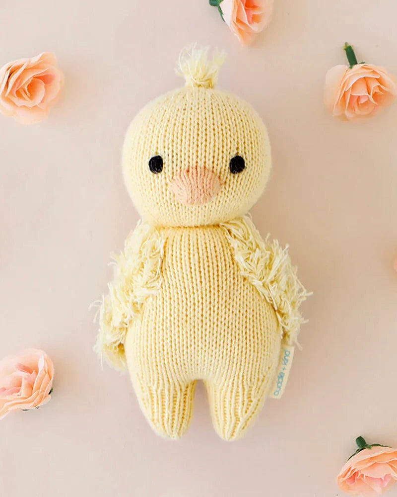 A cute, knitted, yellow Small Easter Basket Set duckling toy with fluffy wings and a tuft on top of its head, surrounded by pink roses on a light background.