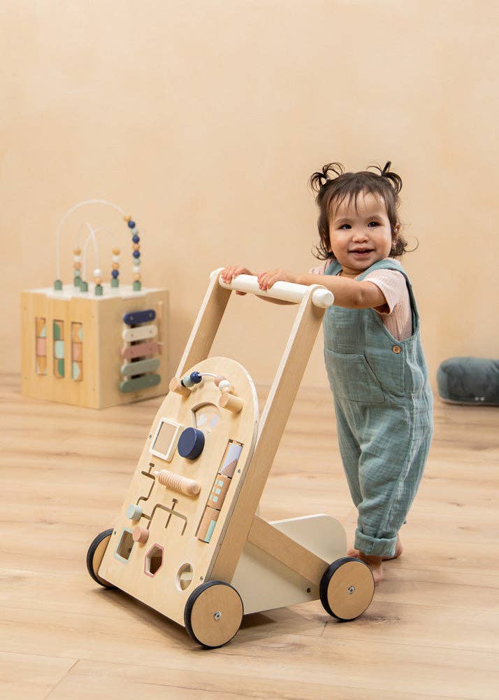 A smiling toddler in blue overalls stands and holds the Wooden Activity Walker. In the background, sensory toys such as a wooden activity cube and a cushion are placed on a light wooden floor.