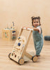 A smiling toddler in blue overalls stands and holds the Wooden Activity Walker. In the background, sensory toys such as a wooden activity cube and a cushion are placed on a light wooden floor.