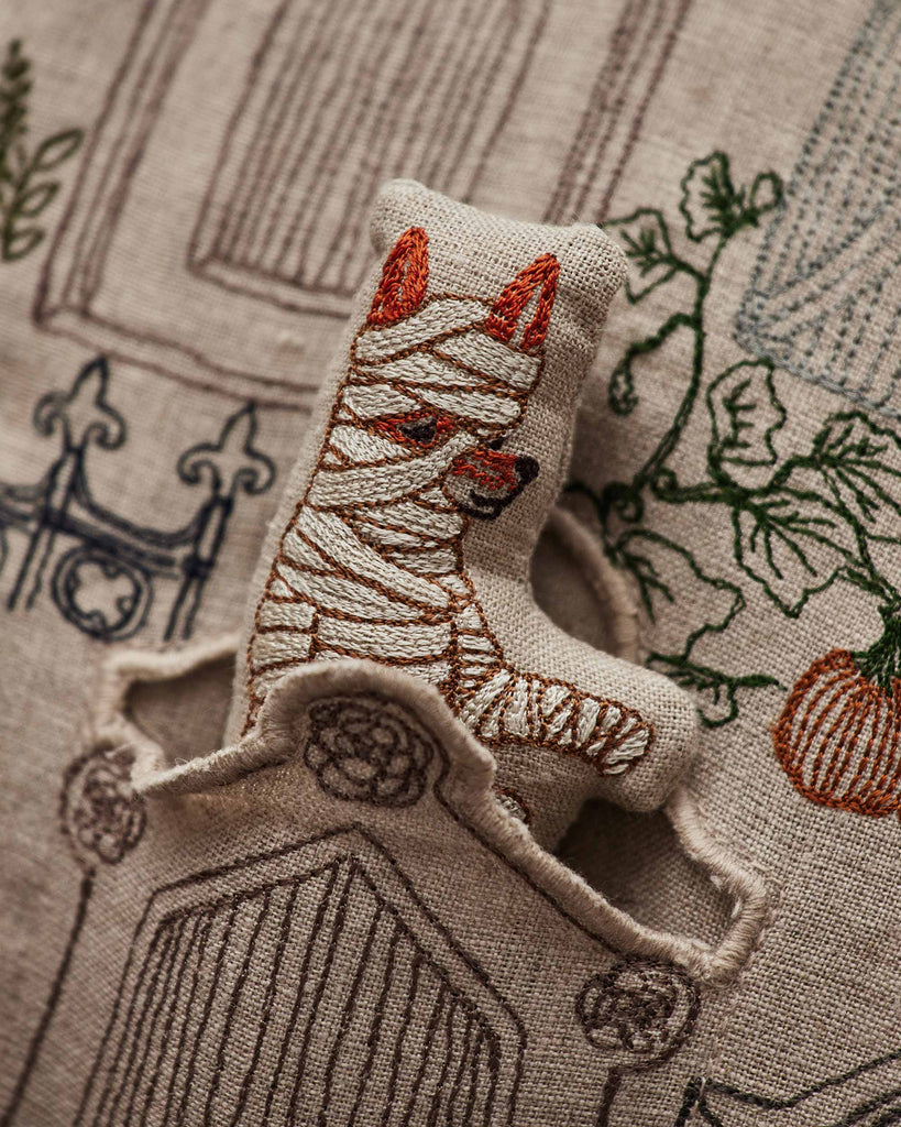 A small, embroidered cat plush wrapped in bandages peeks out from a Coral & Tusk Graveyard Pocket Pillow featuring fabric with garden gate and plant designs.