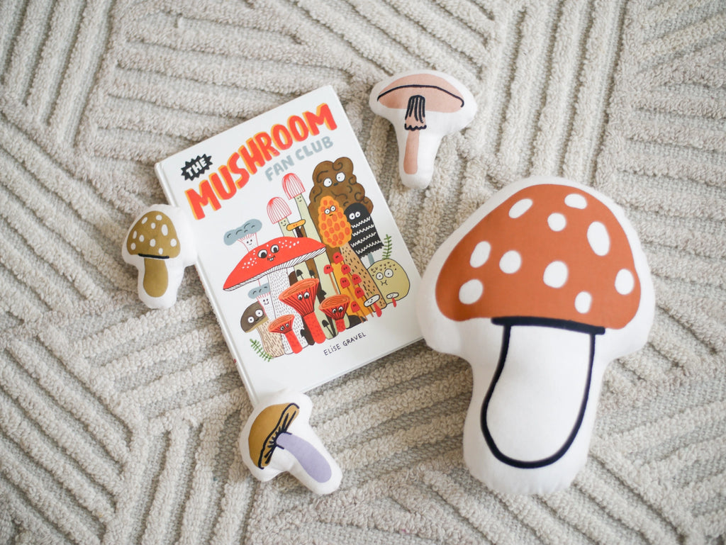 A book titled "Mushroom Fan Club" by Elise Gravel lies on a textured rug, surrounded by Mini Mushroom Baskets in various shapes and colors.