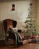 A brown armchair with a green knitted blanket features the Coral & Tusk Christmas Tree Sled Pillow beside a decorated tree with lights and presents underneath, while a black wall sconce glows warmly above.