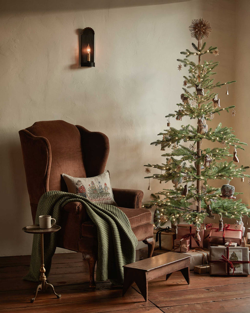 A brown armchair with a green knitted blanket features the Coral & Tusk Christmas Tree Sled Pillow beside a decorated tree with lights and presents underneath, while a black wall sconce glows warmly above.