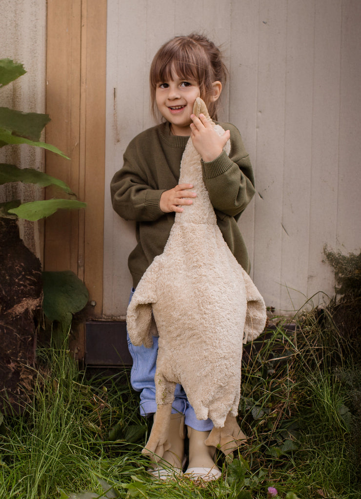 A young child in a green sweater and blue pants smiles while holding a large Senger Naturwelt Cuddly Animal - Linen Goose upright by its neck, standing outside on grass near a wall and wooden door.