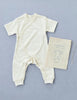 A cream-colored organic cotton Baby Jumpsuit is laid out on a light blue background. Next to it is a matching drawstring bag featuring an illustration of the Baby Jumpsuit and the text "Baby Jumpsuit" and "made with organic cotton.