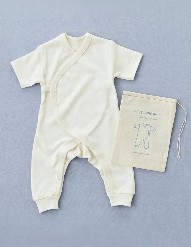 A cream-colored organic cotton Baby Jumpsuit is laid out on a light blue background. Next to it is a matching drawstring bag featuring an illustration of the Baby Jumpsuit and the text "Baby Jumpsuit" and "made with organic cotton.