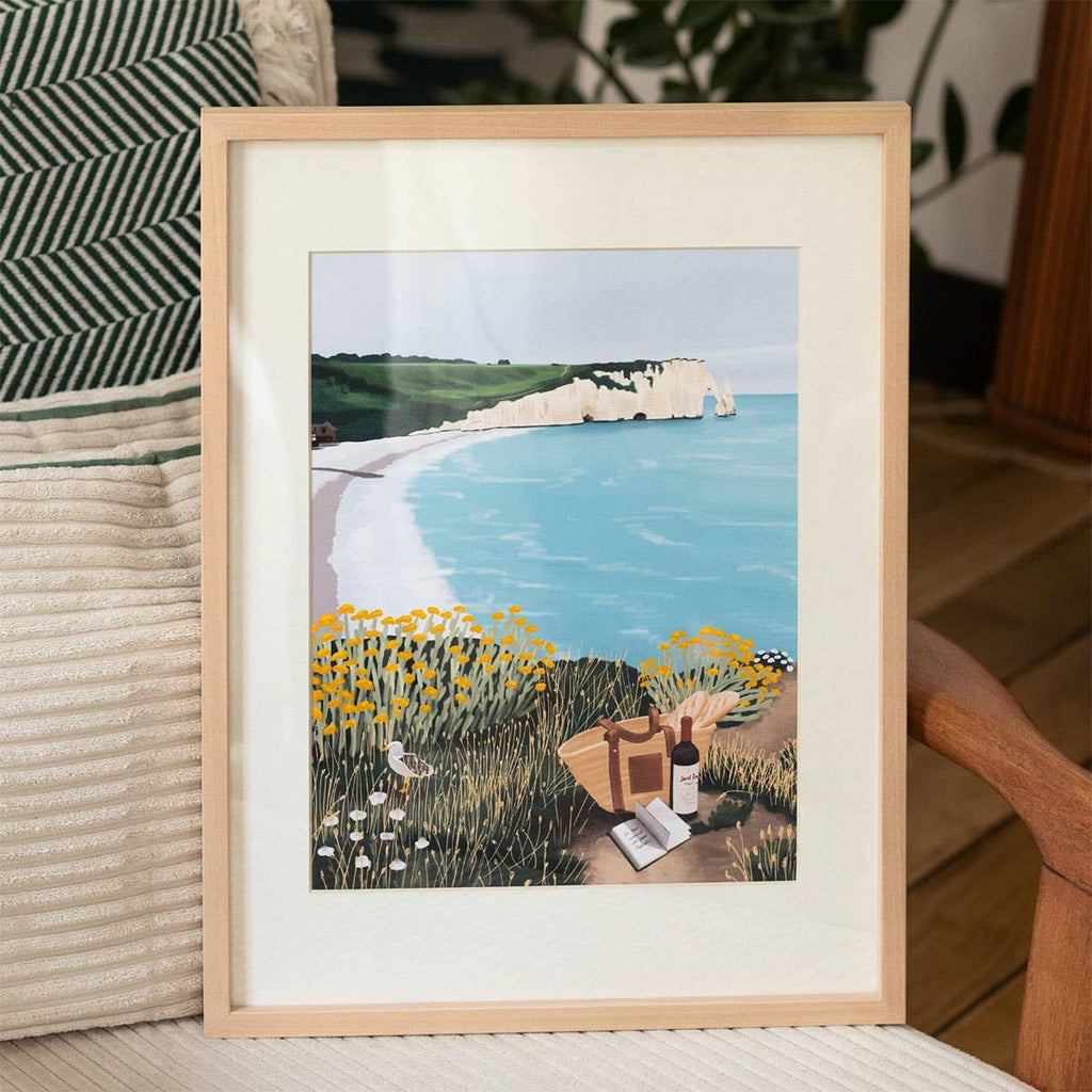 The Paint By Number Kit - Picnic in Étretat by Ananya Bhandari features framed coastal artwork with white cliffs, blue sea, yellow wildflowers, and a picnic setup—displayed on a cushioned chair.