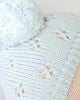 Close-up of the Cuddle + Kind My First Baby (Bluebell) hand-knit light blue doll hat with a large pom-pom and decorative holes, showing part of a person's forehead. Ships in one week.