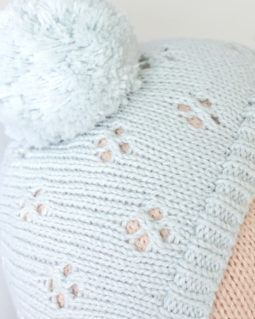 Close-up of the Cuddle + Kind My First Baby (Bluebell) hand-knit light blue doll hat with a large pom-pom and decorative holes, showing part of a person's forehead. Ships in one week.
