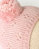 Close-up of the Cuddle + Kind My First Baby (Rose), featuring a pink hand-knit cotton beanie with a large pom-pom and flower-like patterns, shown on a cream mannequin head. Ships in one week.