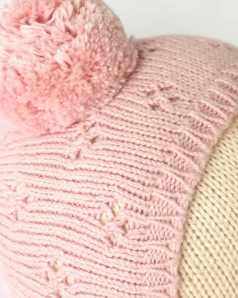 Close-up of the Cuddle + Kind My First Baby (Rose), featuring a pink hand-knit cotton beanie with a large pom-pom and flower-like patterns, shown on a cream mannequin head. Ships in one week.