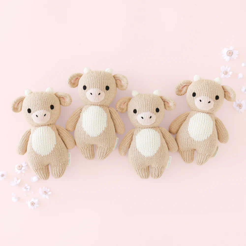 Four Cuddle + Kind Baby Cow toys, crafted from soft Peruvian cotton yarn, display beige bodies, white bellies, and small horns. They are neatly lined up on a light pink background adorned with small white flowers.