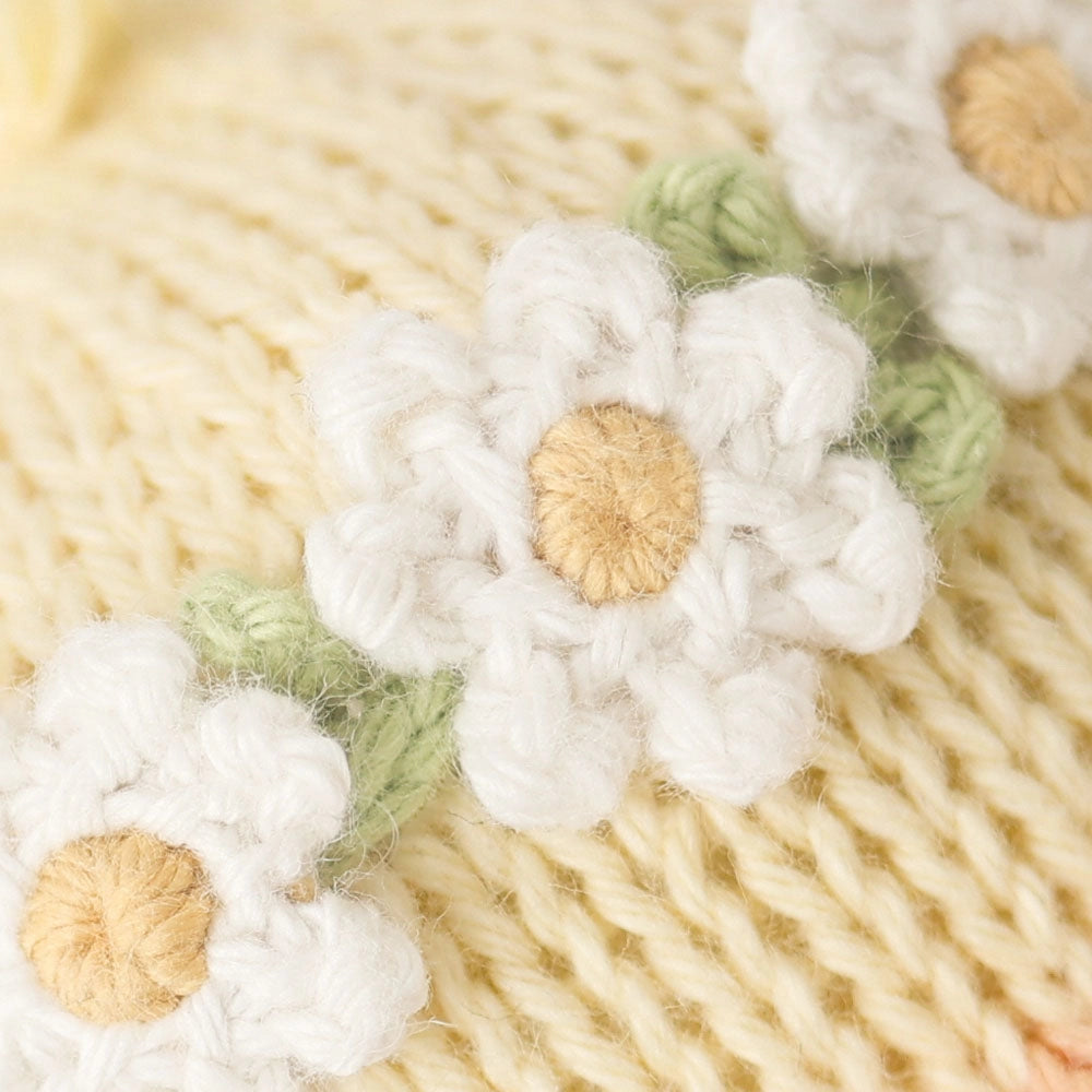 Close-up of the Cuddle + Kind Baby Duckling (Ivory Floral Crown) featuring heirloom-quality, white knitted daisies with tan centers and green leaves on a soft, light yellow Peruvian cotton yarn background.