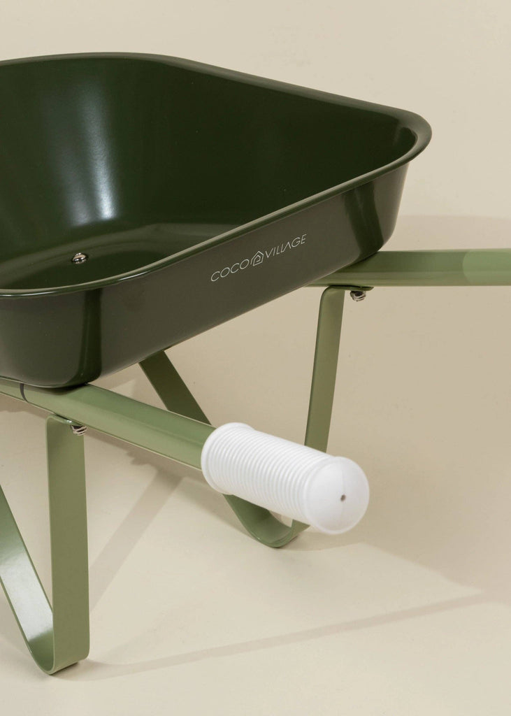 Close-up of the Heritage Wheelbarrow – Liane, a green children's wheelbarrow with a white ribbed handle and part of the frame visible, set against a beige background. "COCO VILLAGE" is printed on the side, making it a charming kids' garden tool.