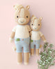 Two Cuddle + Kind Liam the Pony dolls, one large and one small, wear cream shirts and light blue overalls as they lie on a pink background with white flowers. Both have beige manes and friendly faces, lovingly handmade in Peru.