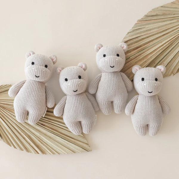 Four Cuddle + Kind Baby Hippo plush toys are neatly lined up on a light surface. In opposite corners, two dried palm leaves, crafted sustainably and fairly, enhance the display with a natural touch.