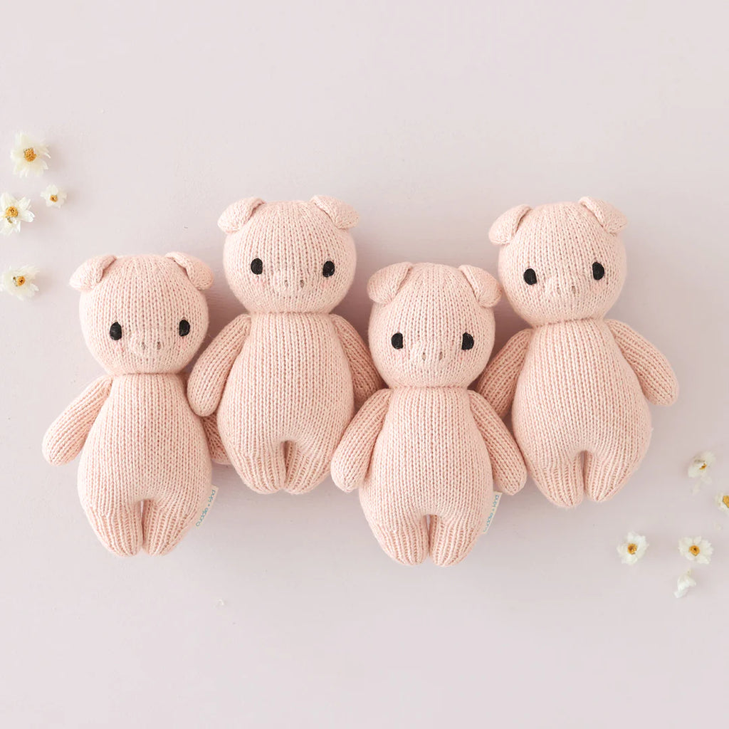 Four Cuddle + Kind Baby Piglet plush toys arranged in a row on a light background, surrounded by small white flowers.