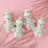 Four adorable hand-knit Cuddle + Kind Baby Zebra dolls in a row on a pink background, surrounded by soft pink palm fronds. Each giraffe has a sweet smile and tufted hair.