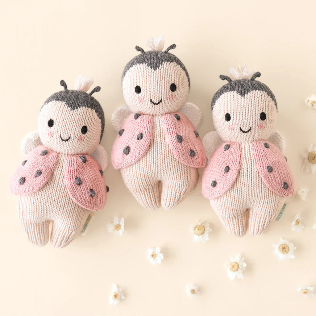 Three Cuddle + Kind Baby Ladybugs with pink wings and smiling faces are arranged on a light surface among small white flowers. Each is hand-knit from natural Peruvian cotton yarn for extra softness.