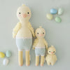 Three Cuddle + Kind Duckling plush toys of varying sizes with pastel-colored eggs beside them on a light grey background. The toys are styled with striped pants and have soft, textured bodies filled with hypoallergenic poly.