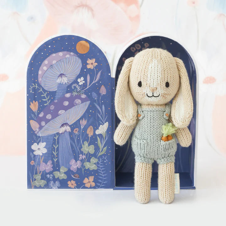 A stuffed bunny toy with long ears, wearing a grey outfit, stands in front of an open decorative box. The box features whimsical illustrations of mushrooms, flowers, and a crescent moon on a navy blue background. Cuddle + Kind Tiny Henry The Bunny is meticulously crafted from 100% cotton for endless cuddles.