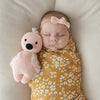 A newborn baby in a yellow floral swaddle sleeps next to a small plush cuddle + kind baby flamingo toy, hand-knit from Peruvian cotton yarn, wearing a light pink bow on her head.