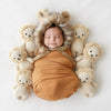 A newborn baby wrapped in an orange swaddle sleeps peacefully, surrounded by six small teddy bears against a white backdrop. The baby wears a hand-knit furry hat with ear flaps made of Cuddle + Kind Baby Lion.