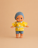 The Minikane Mini Pancha Doll - Basile with Popcorn Painting Outfit features wide eyes and comes dressed in a blue cap, a yellow shirt with white flowers, and blue shorts, standing barefoot on a beige background—perfect for imaginative play.
