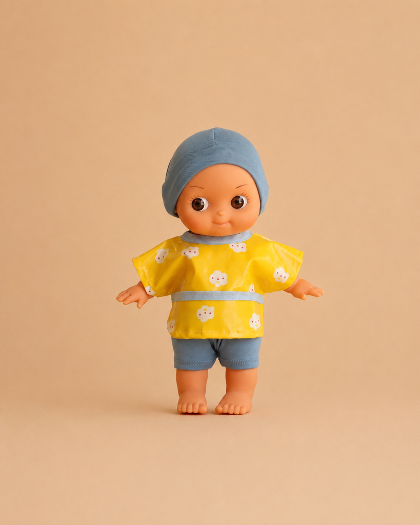 The Minikane Mini Pancha Doll - Basile with Popcorn Painting Outfit features wide eyes and comes dressed in a blue cap, a yellow shirt with white flowers, and blue shorts, standing barefoot on a beige background—perfect for imaginative play.