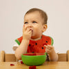 A baby with fruit on their hand sits at a table, eating from the non-toxic Wally the Watermelon Bowl & Spoon Set, wearing a curious expression.